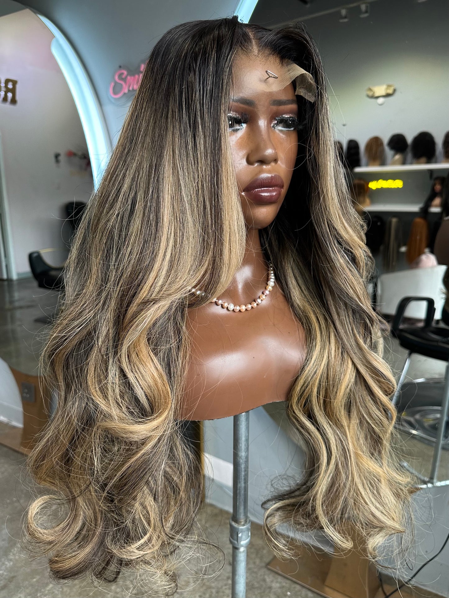 Mannequin head with a long, wavy wig featuring brown and blonde color gradient in a salon setting.