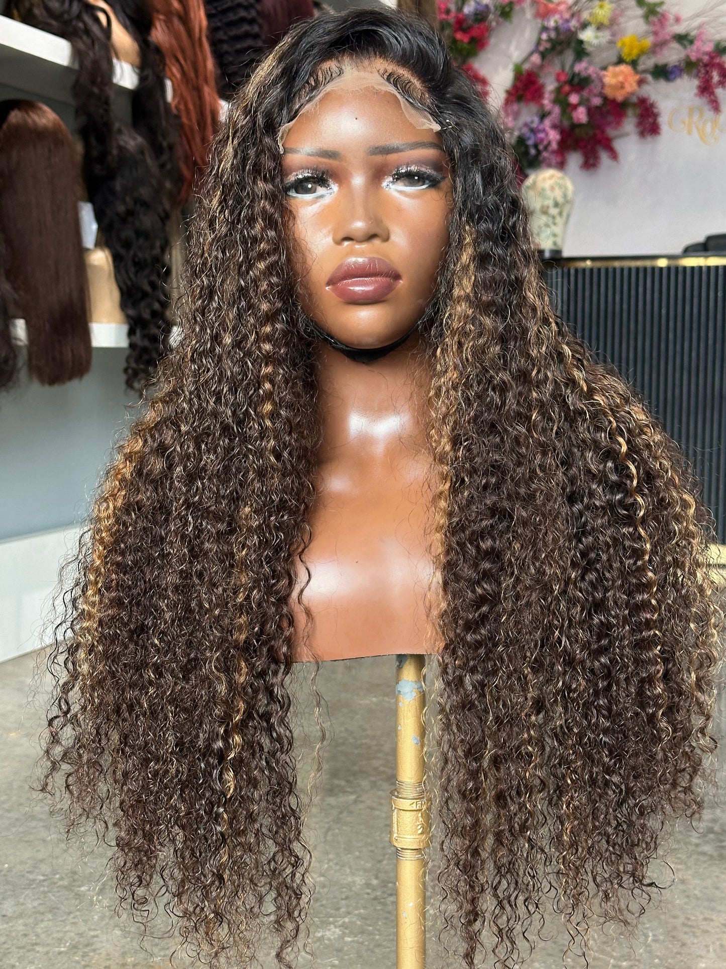 A long, voluminous curly wig displayed on a mannequin head stand in a salon-like setting. The wig has deep, tight curls with subtle warm brown highlights and a black lace frontal part visible at the top.