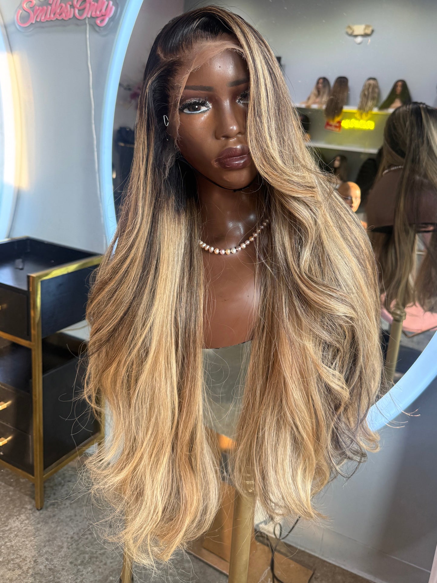 A long wavy wig with dark roots and warm blonde highlights, displayed on a mannequin head inside a salon or boutique setting with a large mirror behind it.